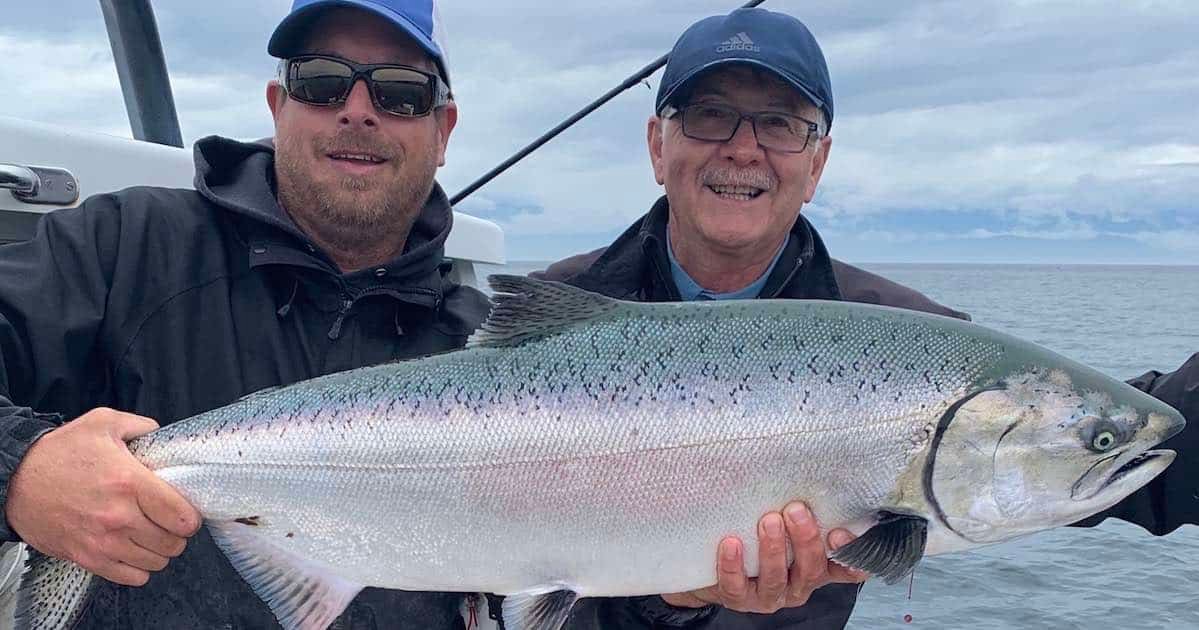 Two men holding a large salmon fish while on a boat in the ocean, with cloudy skies in the background.