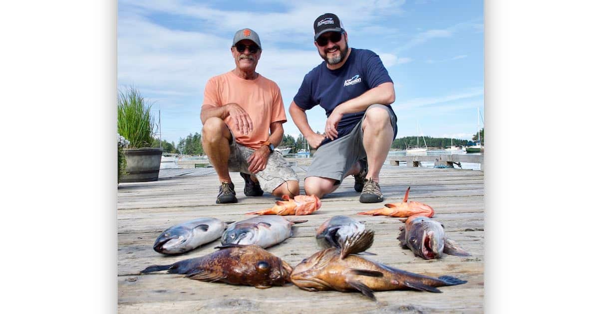 Two men kneeling beside a variety of fish displayed on a wooden dock, with a marina visible in the background.