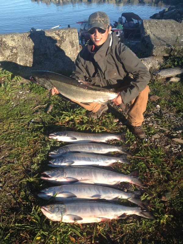 Person holding a large fish while kneeling beside a row of smaller fish on the ground near a body of water.