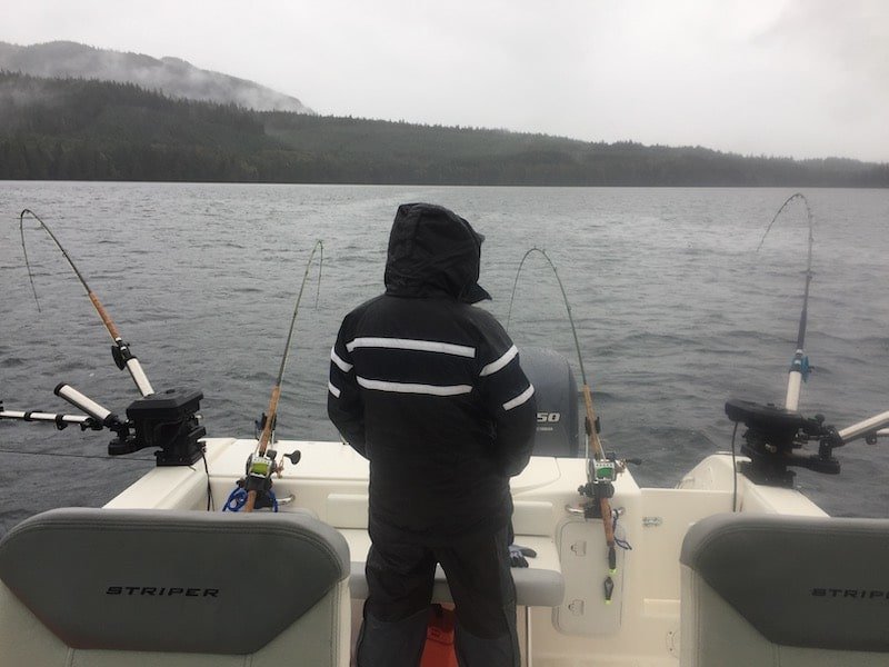 Person in a black and white jacket fishing from a boat with multiple fishing rods set up in the water on a cloudy day.
