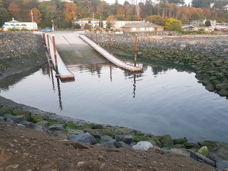Boat ramp leading into calm water, surrounded by rocky shoreline and vegetation, with buildings visible in the background.
