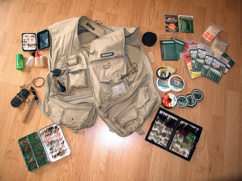 Beige fishing vest laid out with various fishing accessories including flies, tackle boxes, and tools arranged around it.
