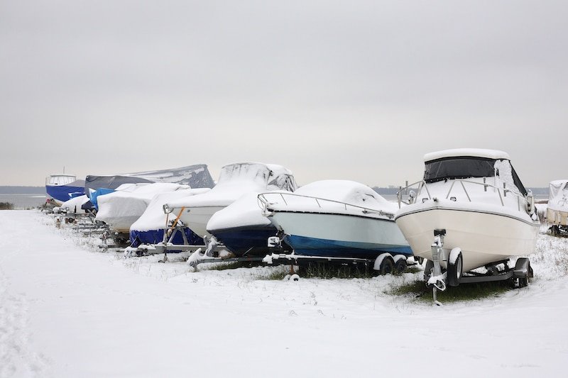 Several boats covered in snow, parked on trailers in a winter setting, showcasing their winter storage condition.