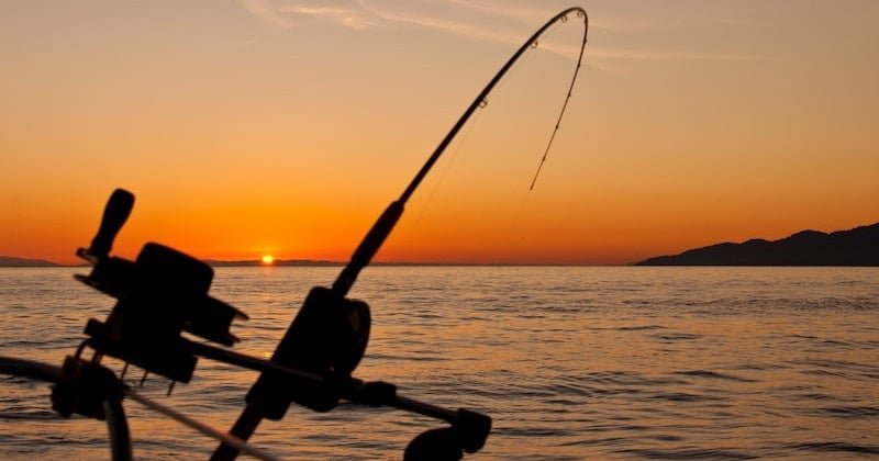 Fishing rod positioned against a sunset backdrop over calm waters, with silhouettes of mountains in the distance.