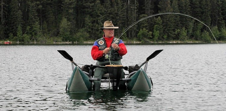 Man fishing from a float tube on a lake, wearing a hat and fishing gear, with a fishing rod bent from a catch.