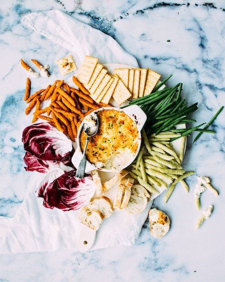 Baked crab dip served in a white dish with assorted crackers, pretzel sticks, and fresh vegetables arranged around it.