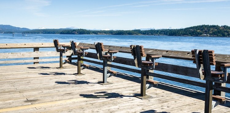 Wooden fishing pier with several benches overlooking a calm body of water and distant mountains under a clear blue sky.