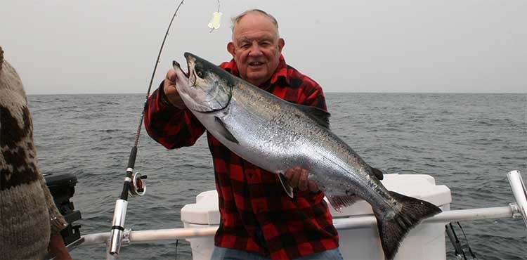 Man holding a large salmon while fishing on a boat in the ocean, wearing a red and black checkered shirt.