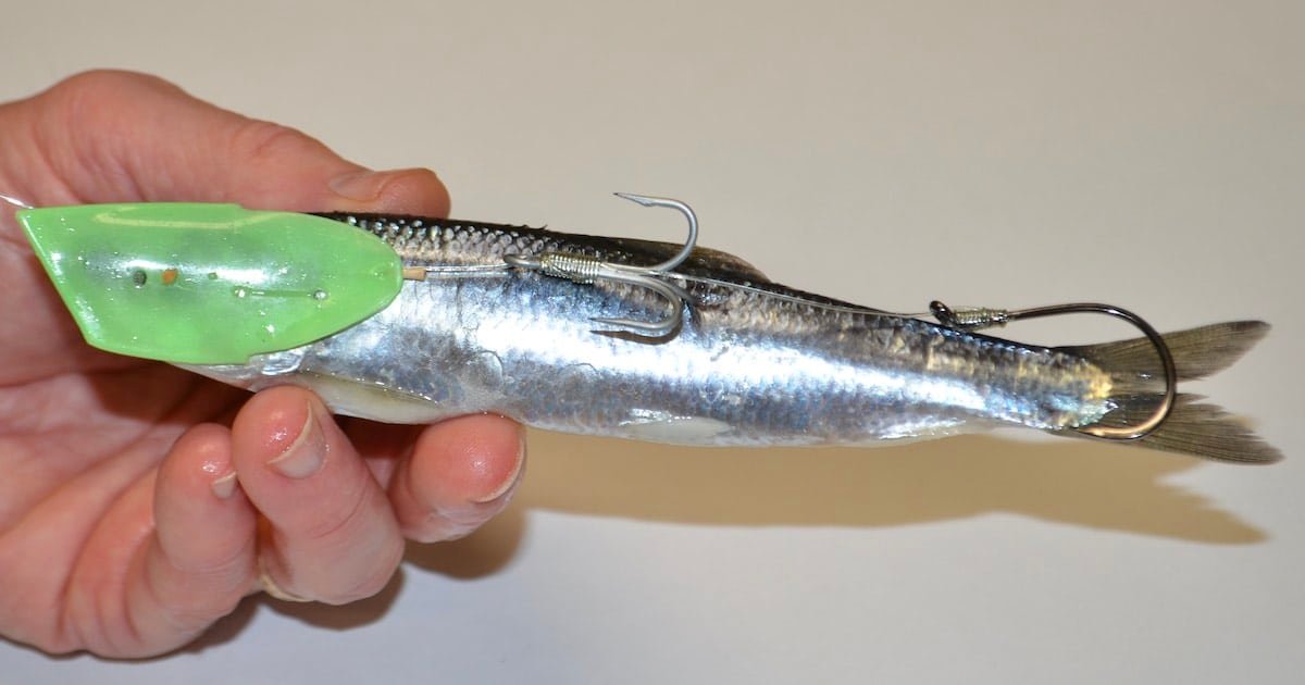 A hand holding a fishing lure that resembles a fish, featuring a green head and two hooks attached to its body.