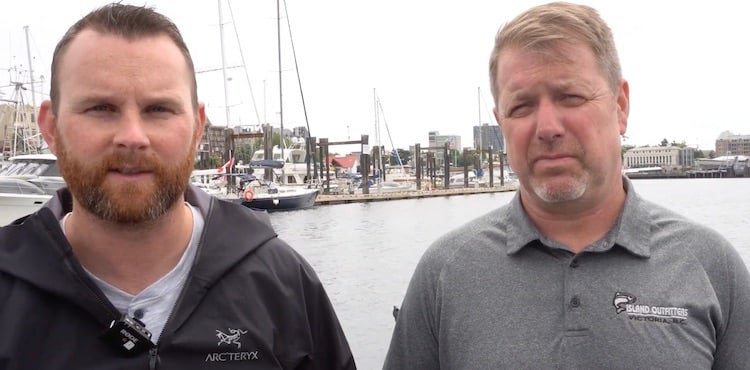 Two men standing by the waterfront with boats in the background, one wearing a black jacket and the other in a gray polo shirt.