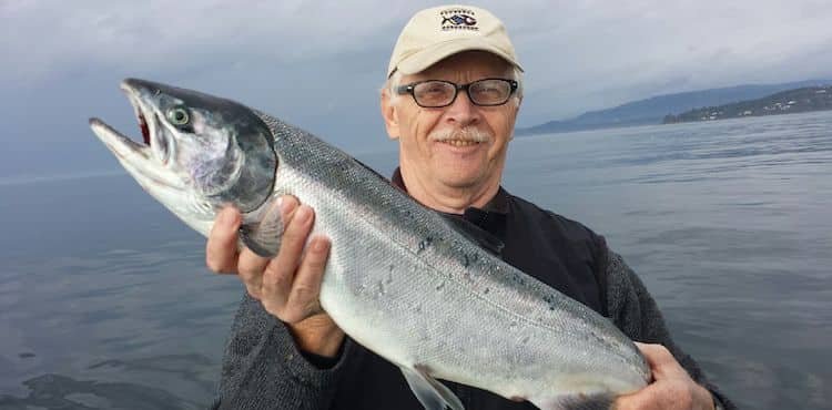 Man holding a coho salmon while standing on a boat, wearing glasses and a cap, with water in the background.