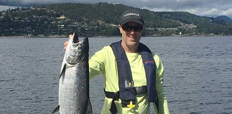 Man holding a large fish while wearing a life jacket and cap, standing on a boat in a body of water.