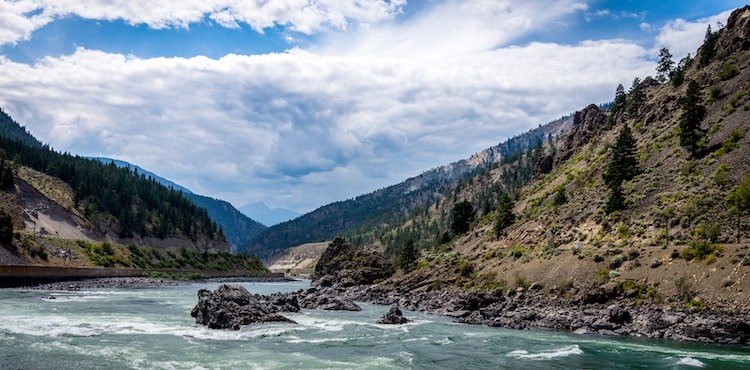 Fraser River flowing through a mountainous landscape with rocky formations and lush greenery along the banks under a cloudy sky.