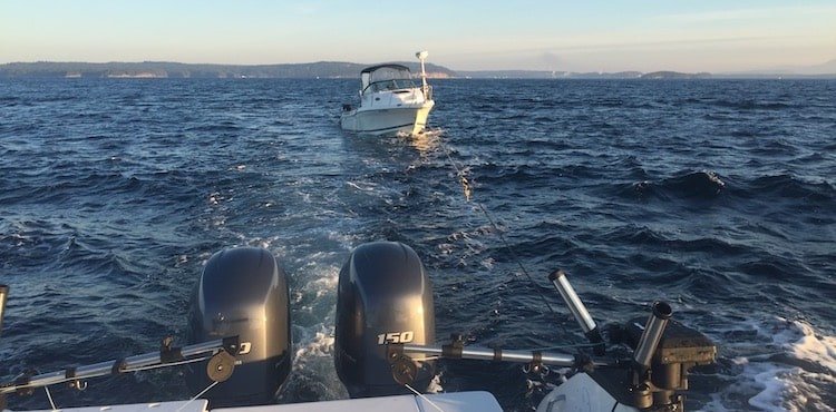 A boat being towed across water, with two outboard motors visible in the foreground. The scene captures waves and a distant shoreline.