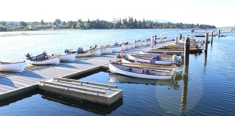 Row of small boats docked along a wooden pier on calm water, with people engaged in various activities nearby.