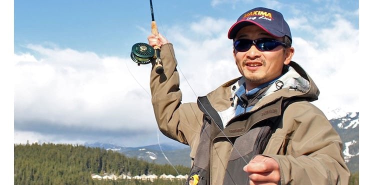 Person holding a fishing rod with a fly reel, wearing sunglasses and a cap, dressed in outdoor fishing attire.