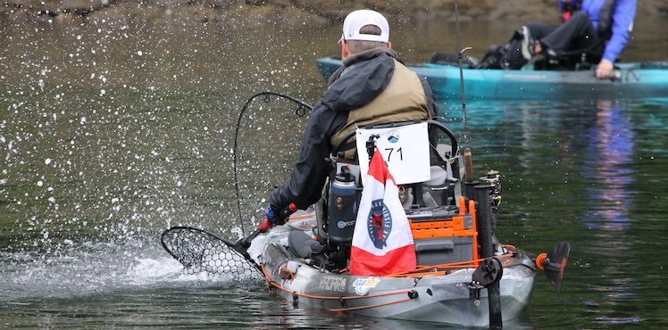 Person in a kayak with a fishing net and a flag, paddling on calm water during a fishing event.