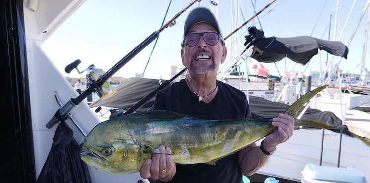Man holding a large mahi-mahi fish while standing on a boat, with fishing rods visible in the background.