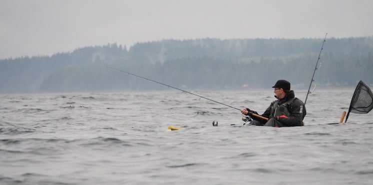 Person fishing from a kayak in open water, wearing a hat and holding a fishing rod, with a net nearby.