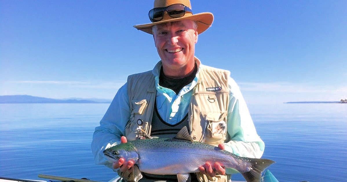 Man wearing a hat and sunglasses holding a Coho salmon while on a boat in calm waters.