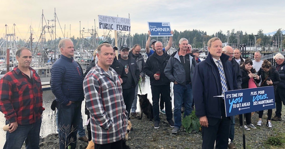Group of individuals gathered near a marina, some holding signs supporting public fishery and a political candidate, with boats in the background.