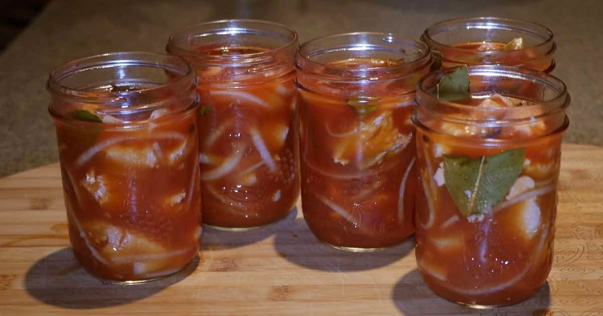 Five jars of pickled salmon with visible spices and ingredients, arranged on a wooden surface.
