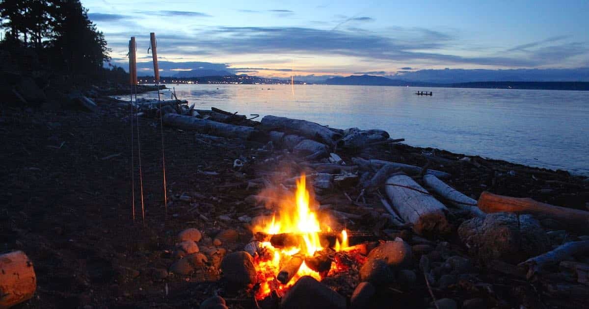 Campfire burning on a beach at dusk, surrounded by logs and stones, with a calm water view in the background.