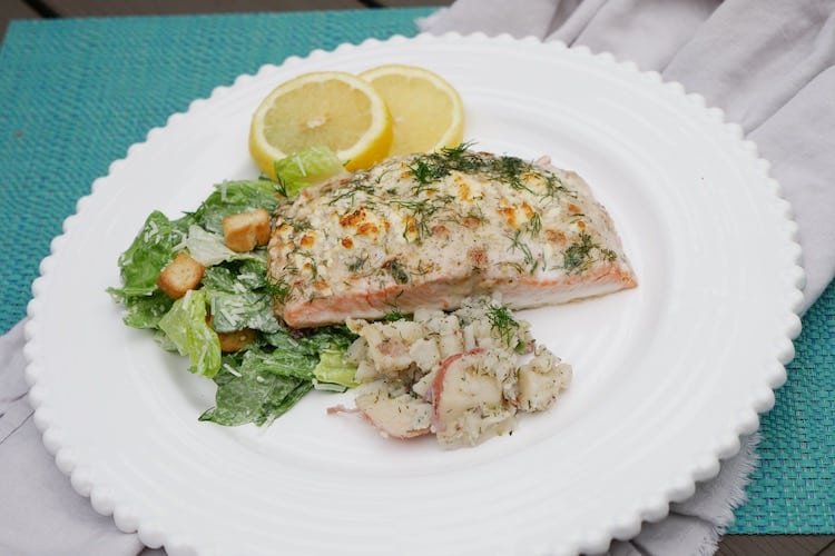 Plate of baked salmon garnished with dill, served with lemon slices, a side salad, and potato salad.