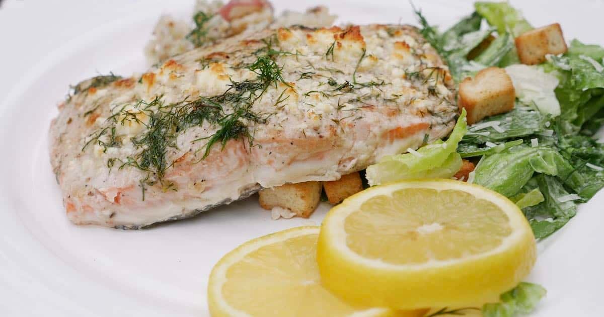 Baked salmon fillet topped with herbs and served with a side salad and lemon slices on a white plate.