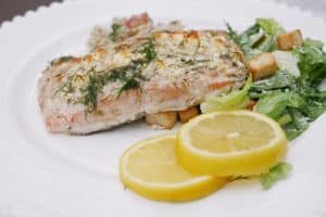 Cooked salmon fillet garnished with dill, served with a side of lettuce and croutons, accompanied by lemon slices.