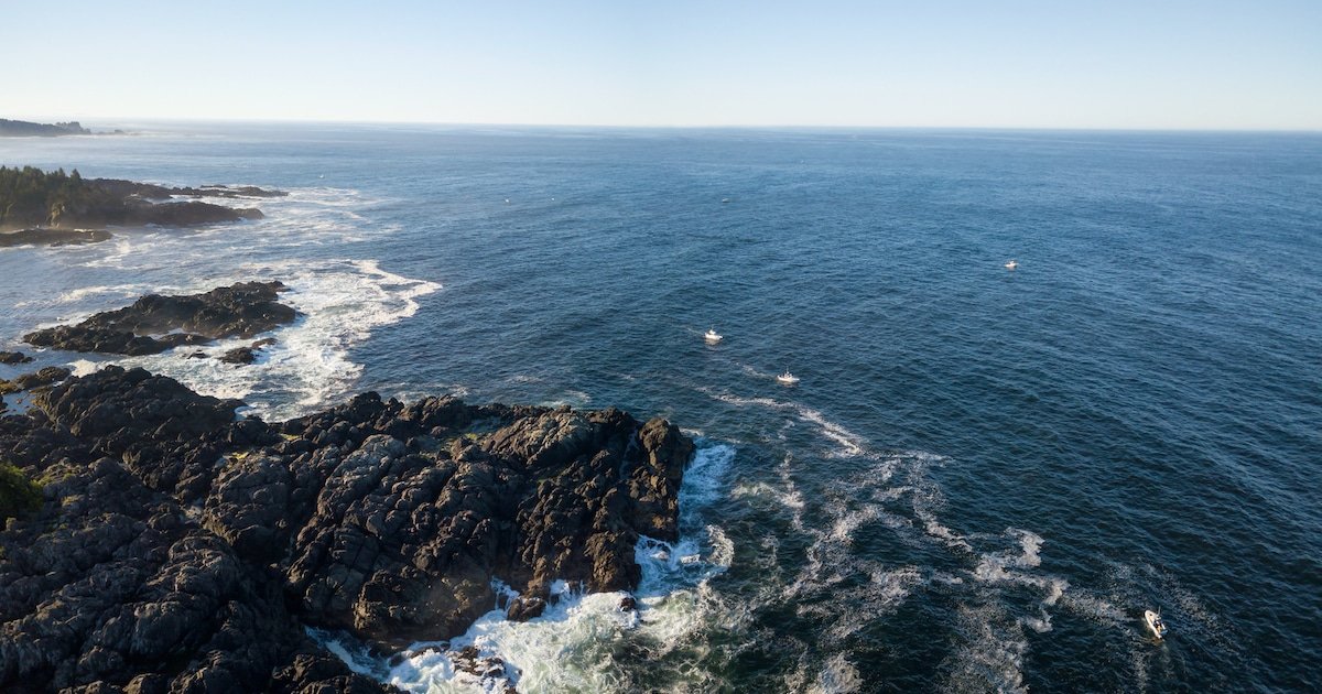Aerial view of rocky coastline with waves crashing against the shore and small boats visible in the ocean.