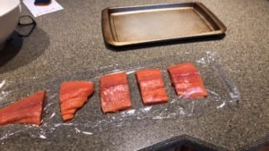 Five salmon fillets arranged on a sheet of plastic wrap on a countertop, with a baking tray visible in the background.