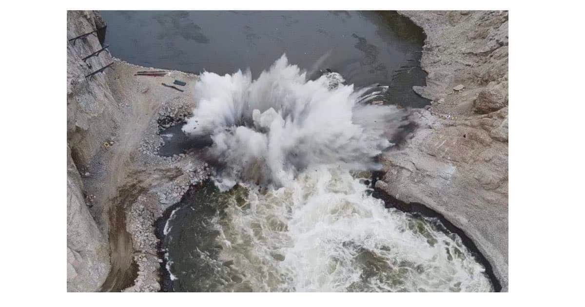 Controlled explosion creating a large plume of water and debris in a river, with rocky banks visible on either side.