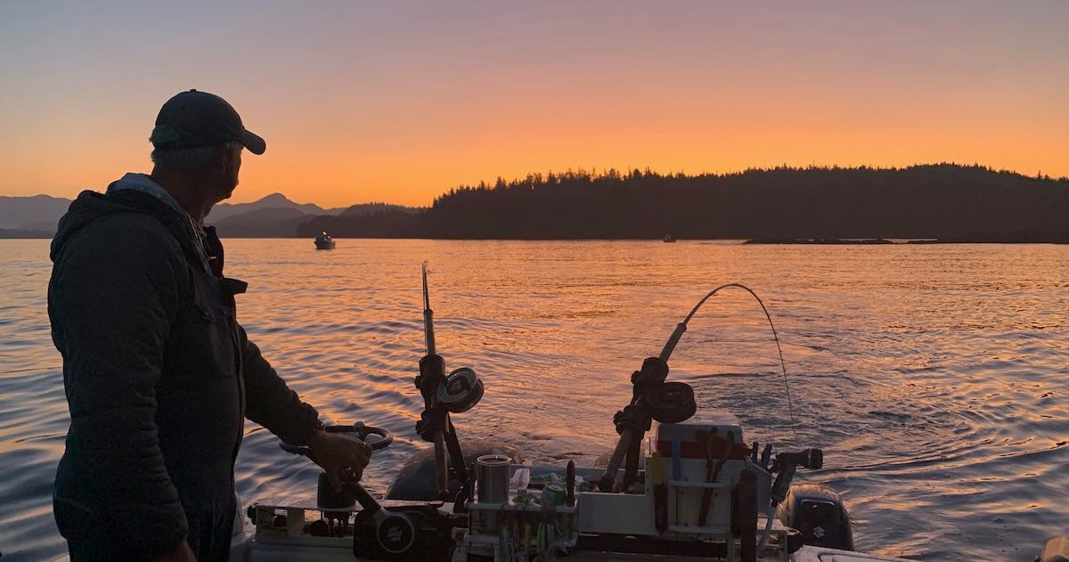 Person operating a fishing boat at sunset on water, with fishing gear visible and silhouettes of other boats in the background.