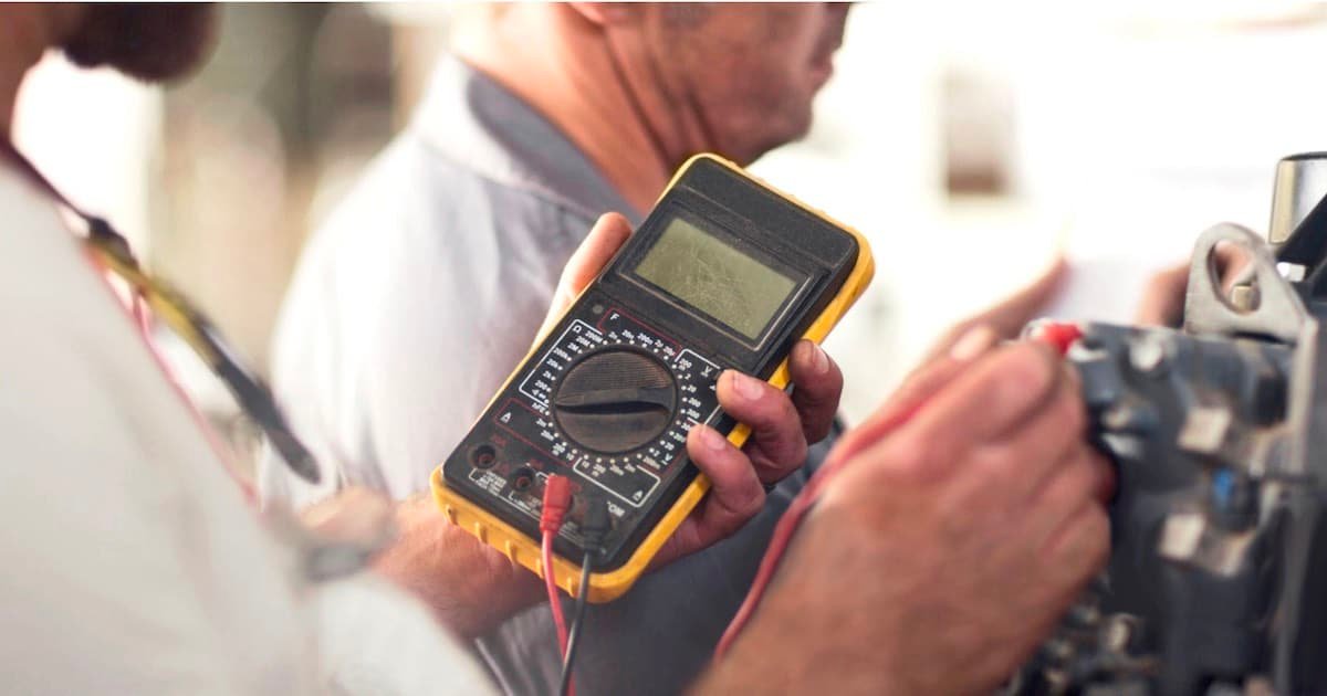 Digital multimeter held by a person, displaying readings while testing electrical components in a workshop setting.