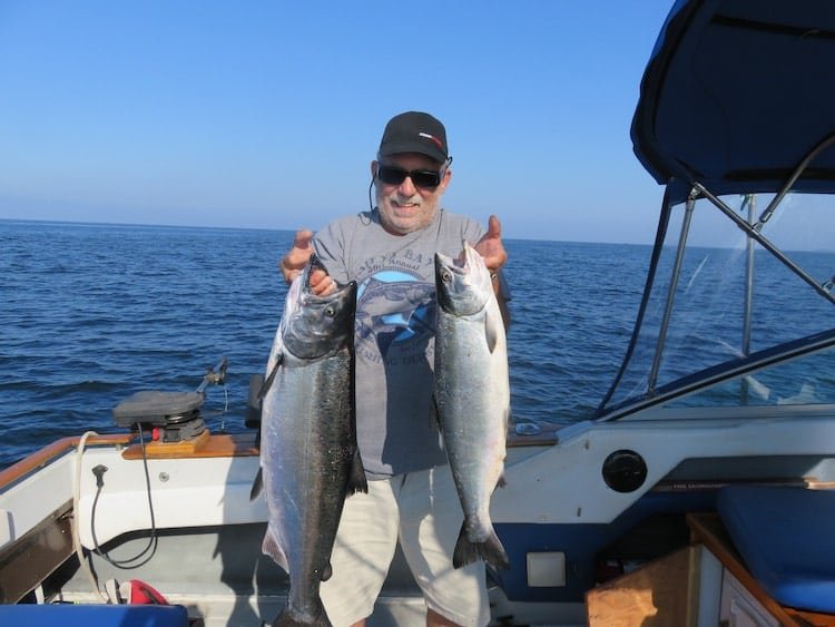Man holding two large fish while standing on a boat in the ocean, wearing a cap and sunglasses.