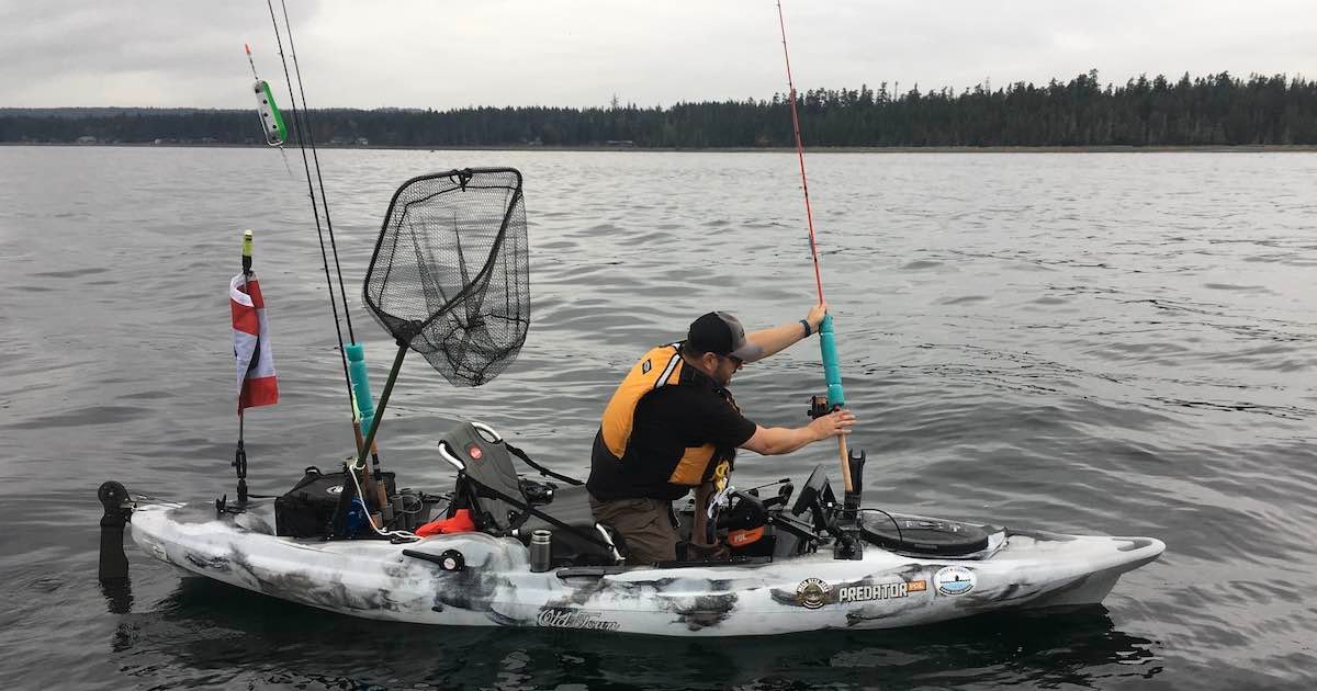 Person in a kayak adjusting fishing gear while on calm water, with fishing rods and a net visible on the kayak.