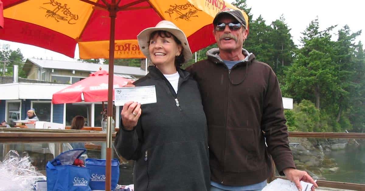 Woman holding a winning cheque for the largest Chinook in a fishing derby, standing next to a man under an umbrella.