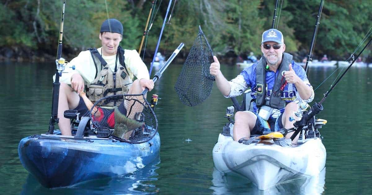 Two individuals sitting in kayaks on water, one holding a fishing net and both giving a thumbs up.