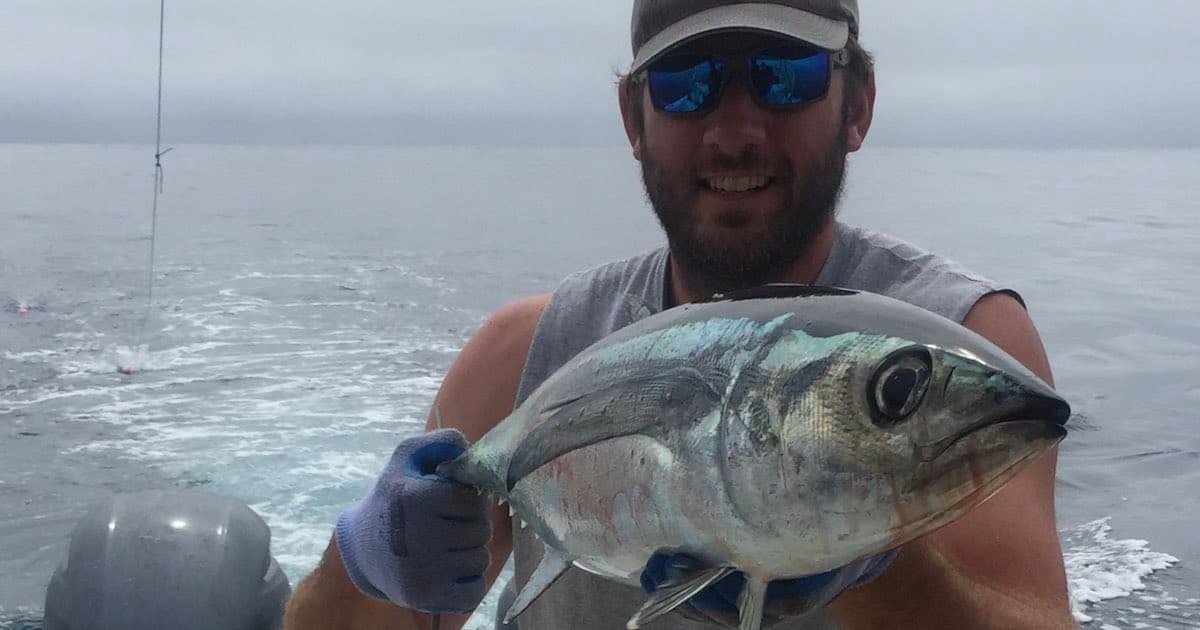 Man holding a tuna fish while wearing sunglasses and gloves, with water visible in the background.