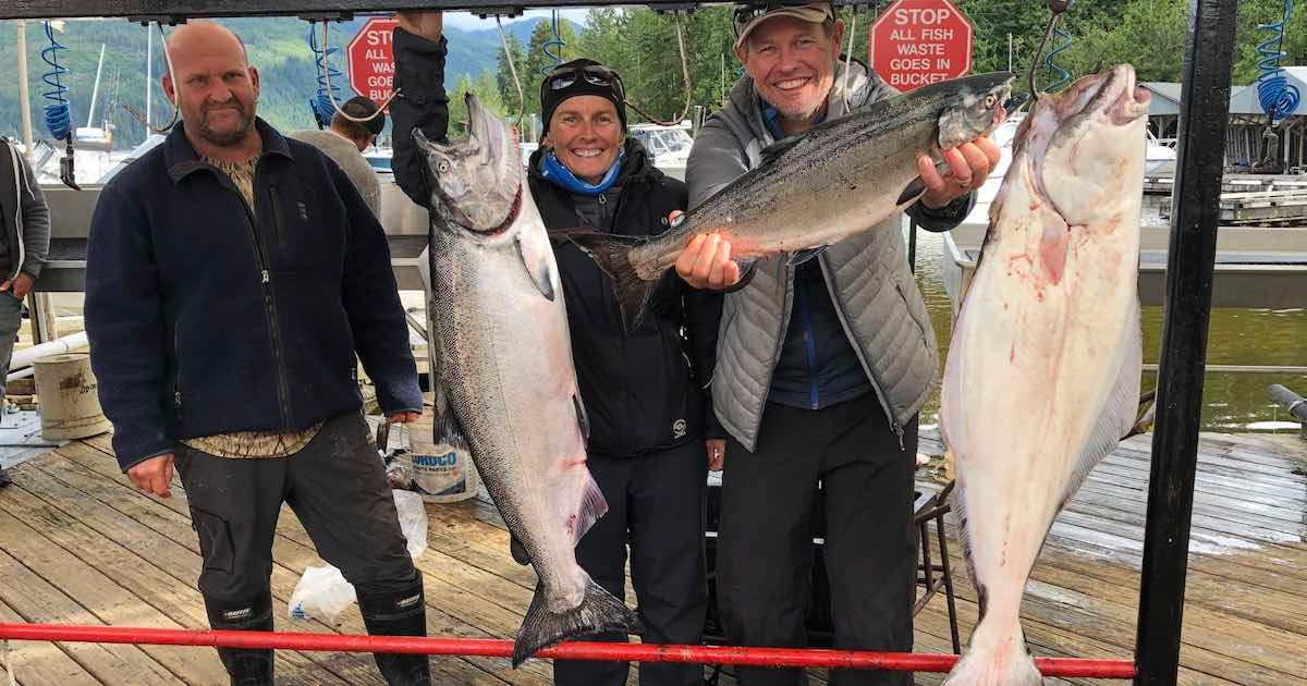 Three individuals holding large fish, including salmon and halibut, displayed on a rack at a fishing dock.