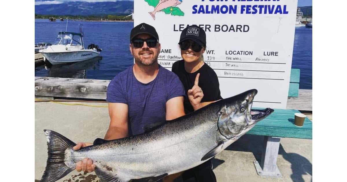 Man and woman holding a large Chinook salmon at a fishing festival, with a leaderboard in the background.