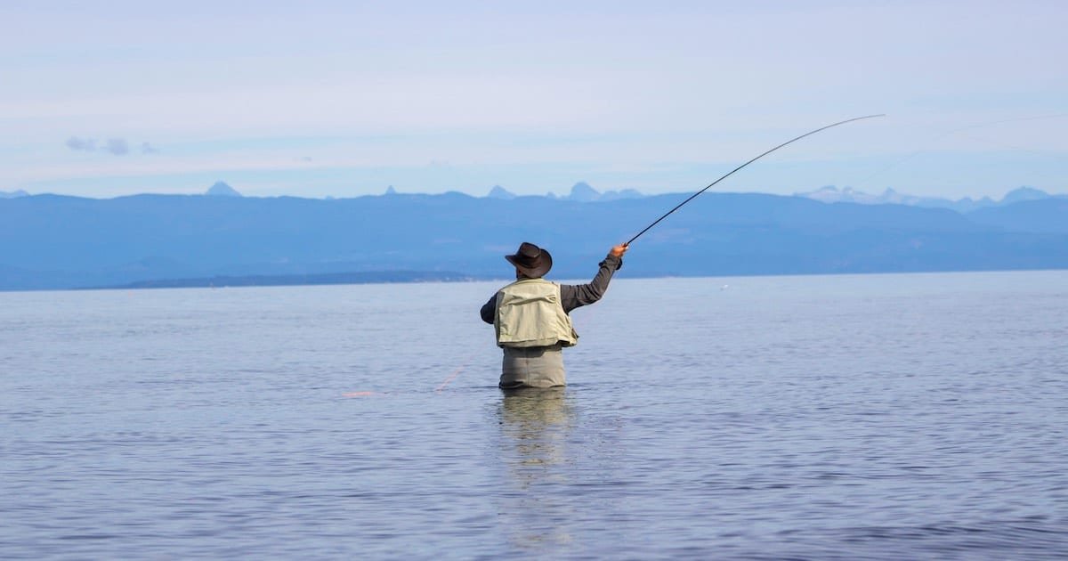 Person casting a fishing line while standing in shallow water, wearing a hat and fishing gear, with mountains visible in the background.