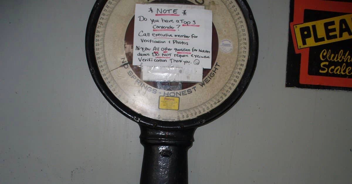 A vintage mechanical scale with a circular dial and a note attached, indicating instructions for verification and photo submission.
