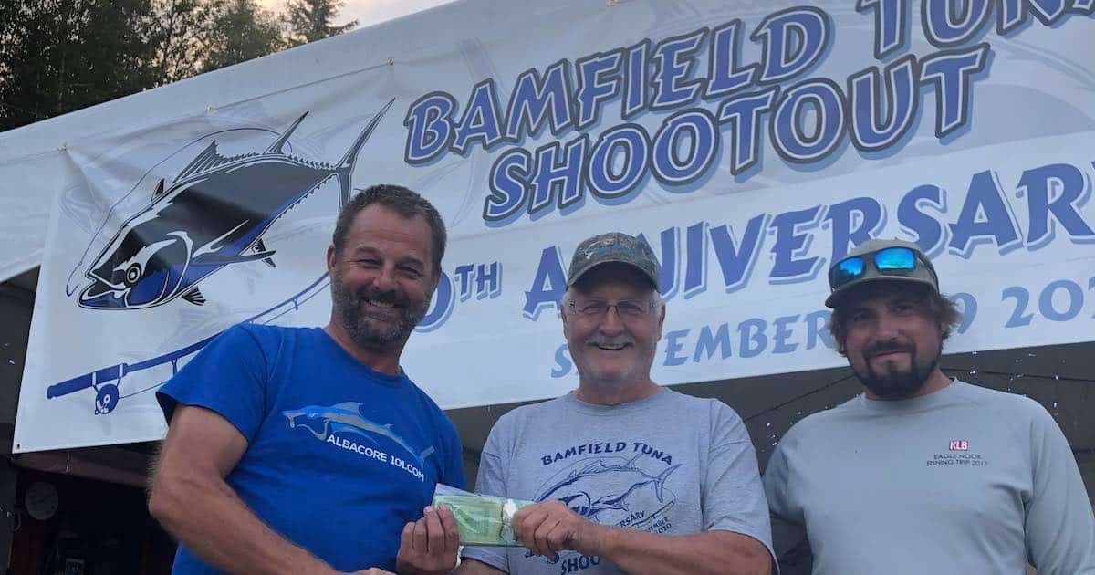 Three men holding a cash prize at the Bamfield Tuna Shootout event, celebrating its anniversary with a banner in the background.