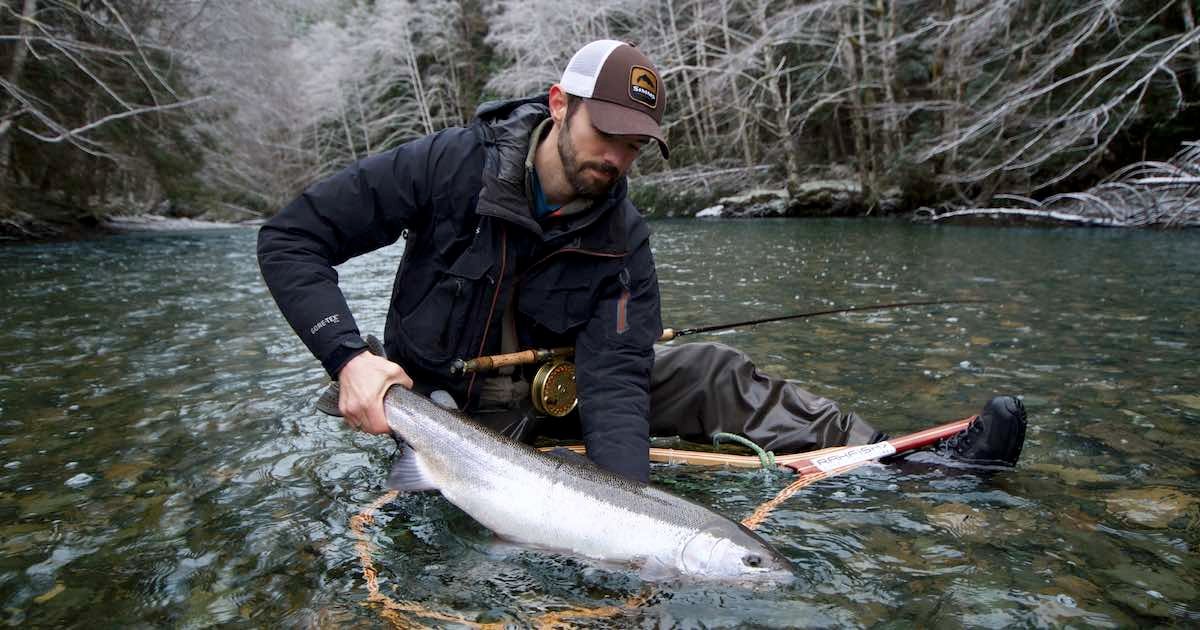 Angler holding a large steelhead trout while seated in a river, wearing fishing gear and a cap.