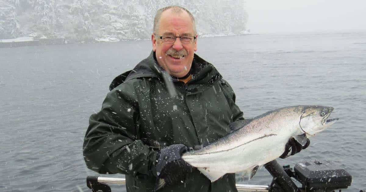 Man holding a large fish while wearing a green jacket and gloves in snowy conditions near water.