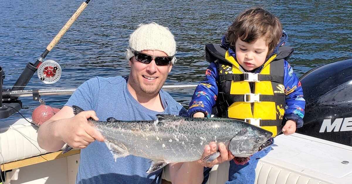 Man holding a fish while a child stands beside him on a boat, both wearing casual clothing and a life jacket.