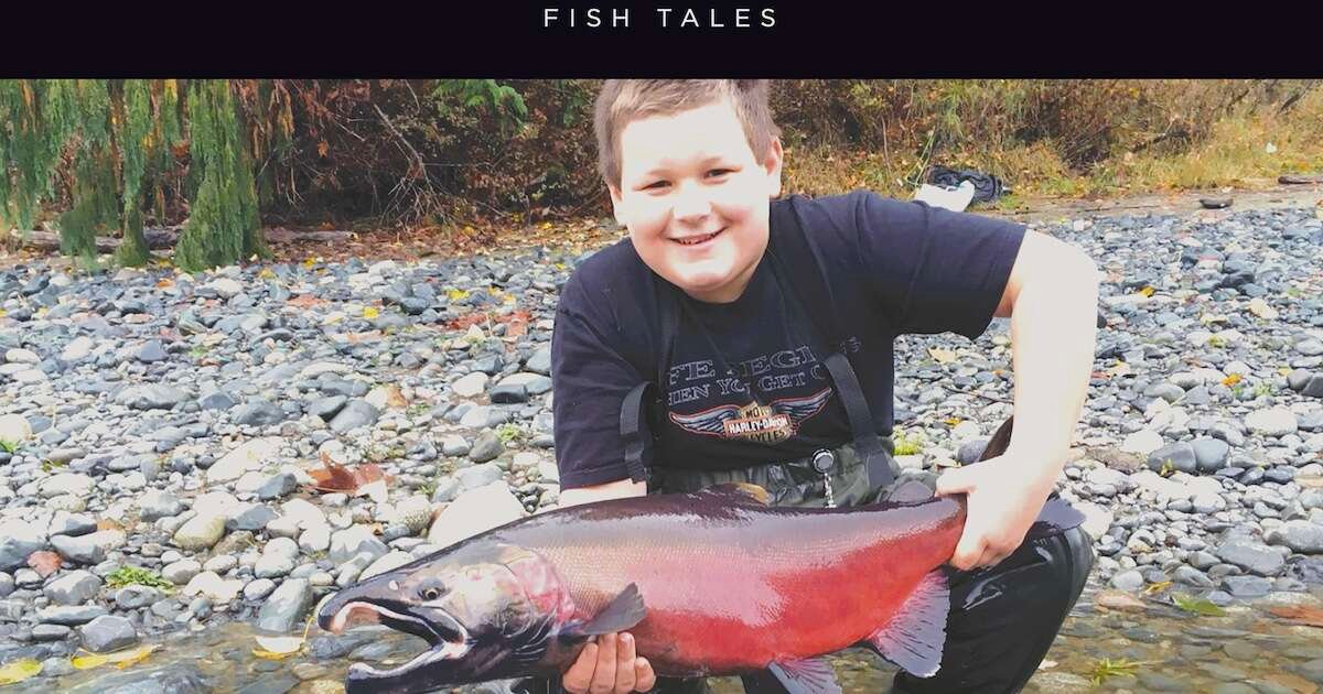 Young boy holding a large salmon fish, smiling while standing on a rocky riverbank.