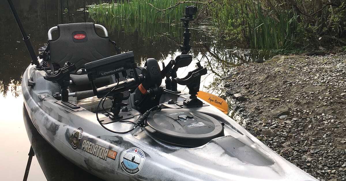 A kayak equipped with various fishing accessories, including a fishing rod holder and a camera mount, positioned near a waterway.